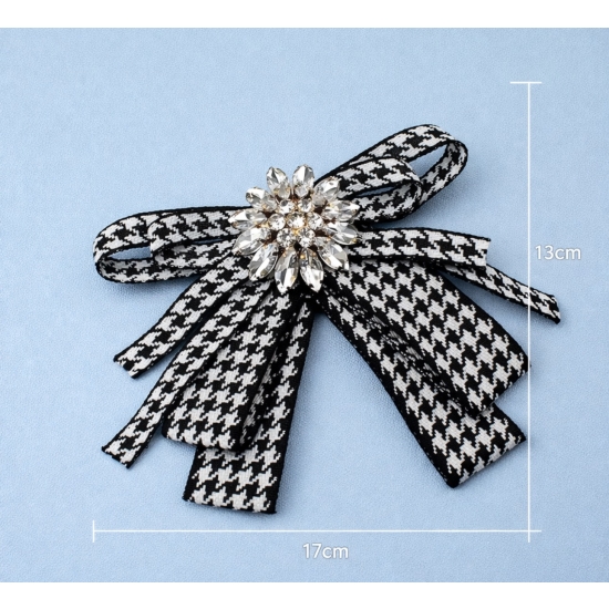 Stylowa broszka w formie kokardy w klasyczny wzór pepitki, ozdobiona efektownym kryształowym środkiem – elegancki detal, który przyciąga spojrzenia.